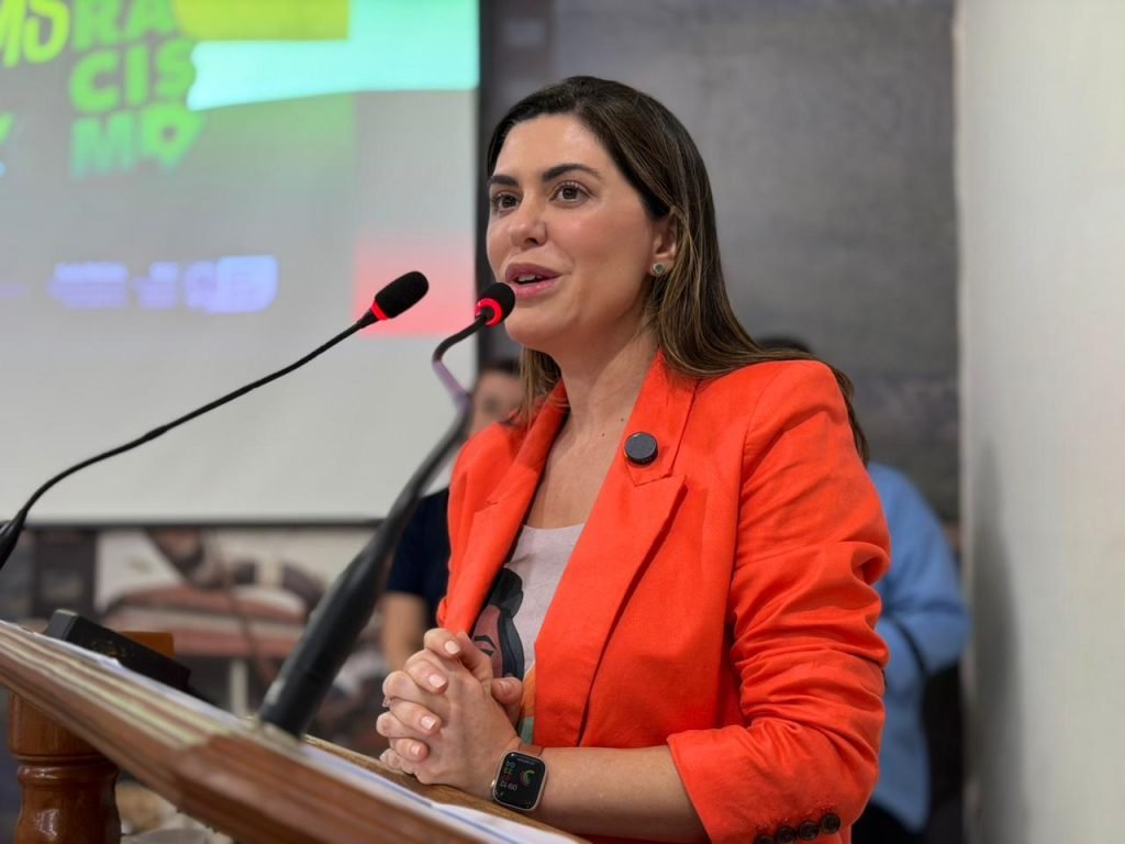Uma mulher branca com cabelo castanho, vestindo um blazer laranja vibrante, em um pódio. Ela está falando em dois microfones, com as mãos unidas e uma tela de projeção com a frase "MS sem Racismo" desfocada ao fundo.
